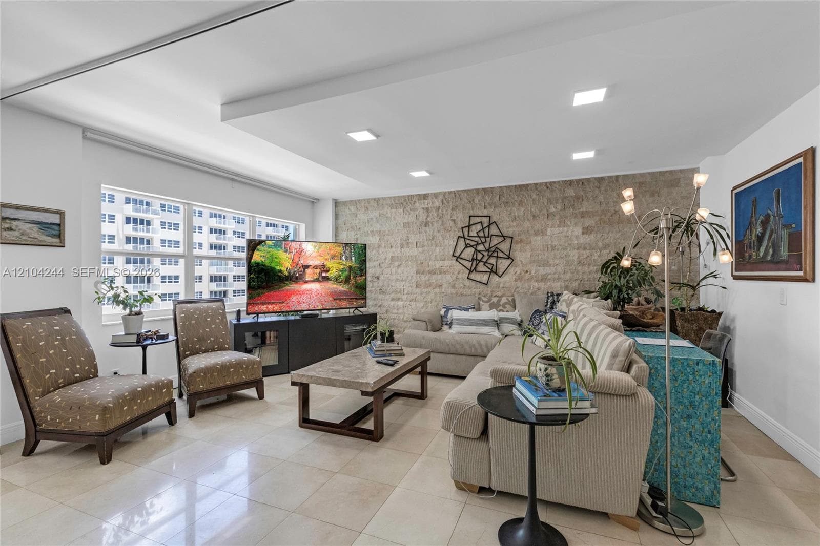 Living area featuring a textured stone accent wall, large-format tile flooring, a recessed tray ceiling with integrated lighting, and expansive windows