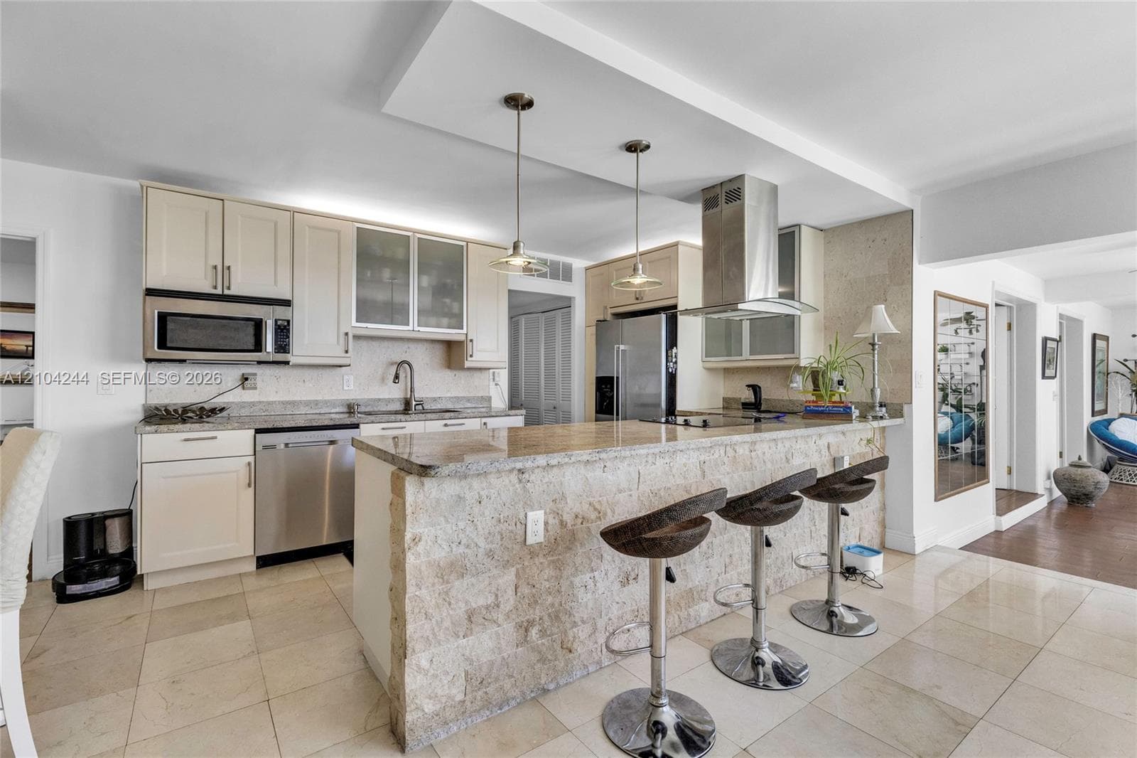 Kitchen featuring stone-clad breakfast bar, stone countertops, stainless steel appliances, cabinet-fronted upper storage with glass inserts, and two pendant lights suspended from a drop ceiling
