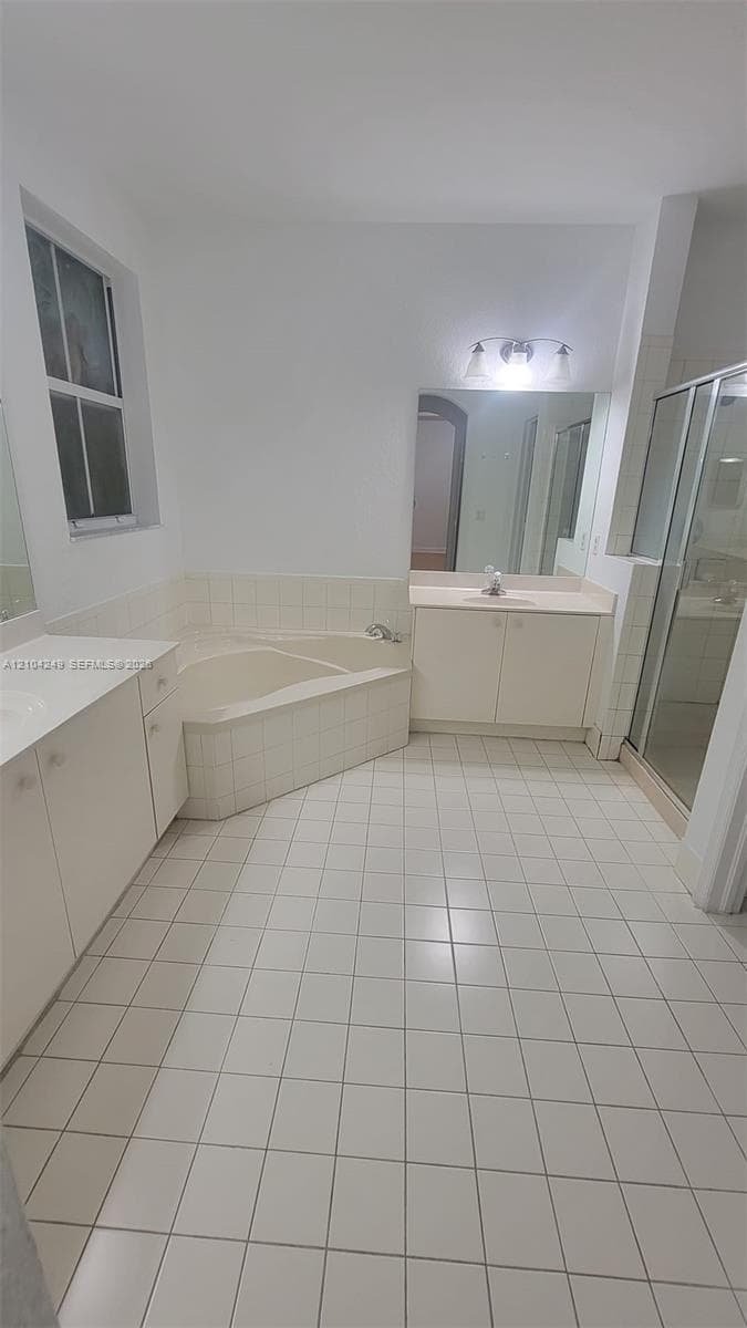 Master Bathroom featuring dual separate vanities, a tiled soaking tub, a glass-enclosed shower, and light-toned ceramic tile flooring