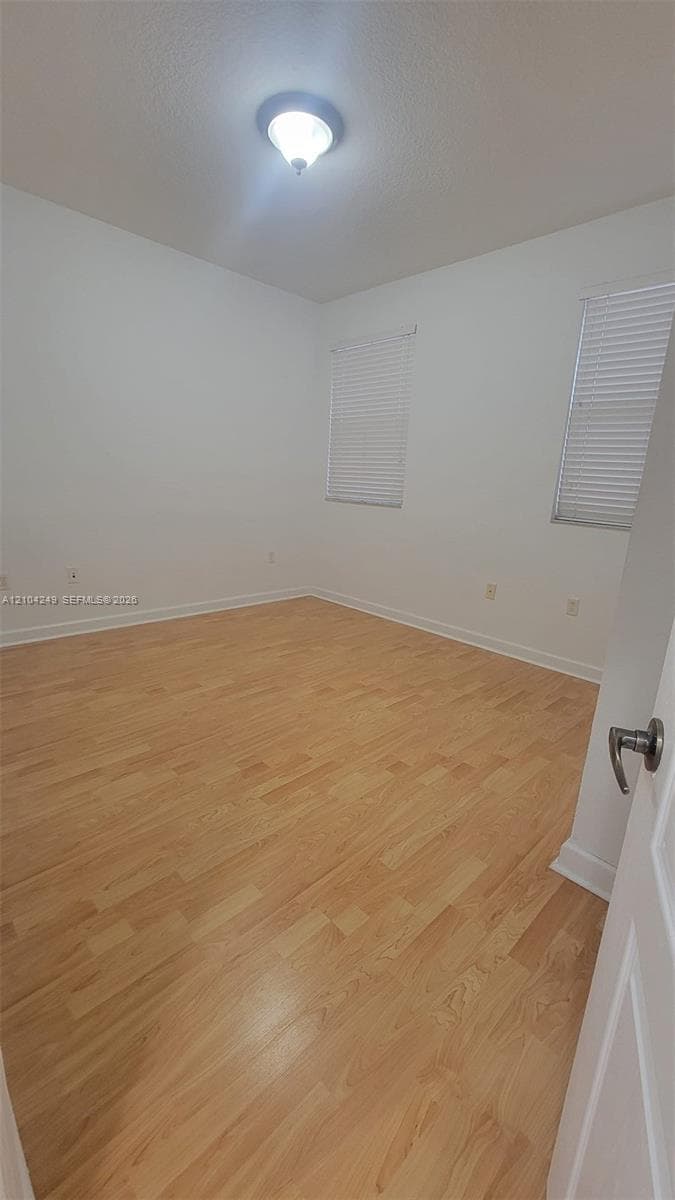 Room interior featuring light-tone wood-finish flooring, neutral walls, two windows with installed horizontal blinds, and a central ceiling-mounted light fixture