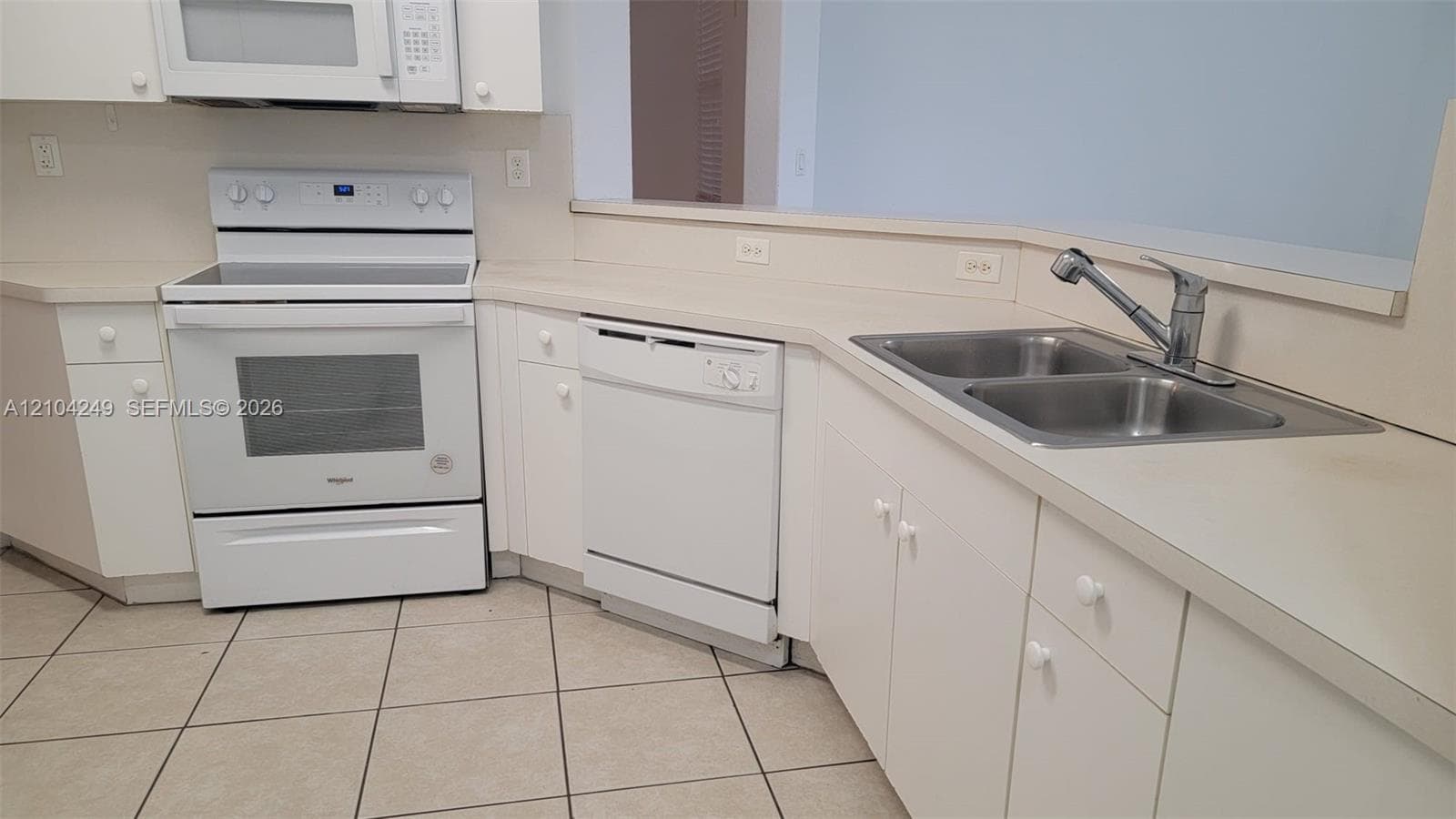 Kitchen featuring white cabinetry, solid-surface countertops, dual-basin stainless steel sink with pull-down faucet, white built-in appliances, and tile flooring