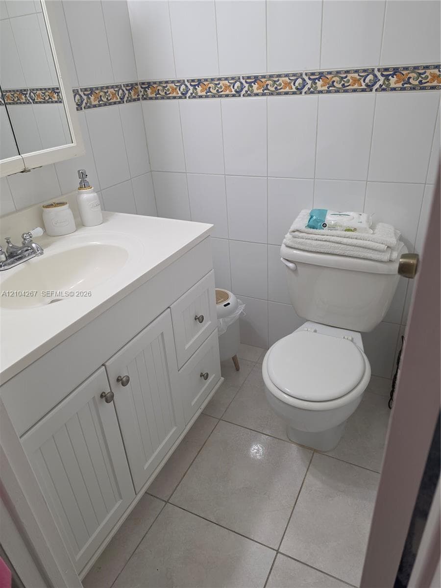 White vanity with beadboard cabinet doors, integrated sink, tiled wall surround featuring a decorative accent border, and ceramic tile flooring