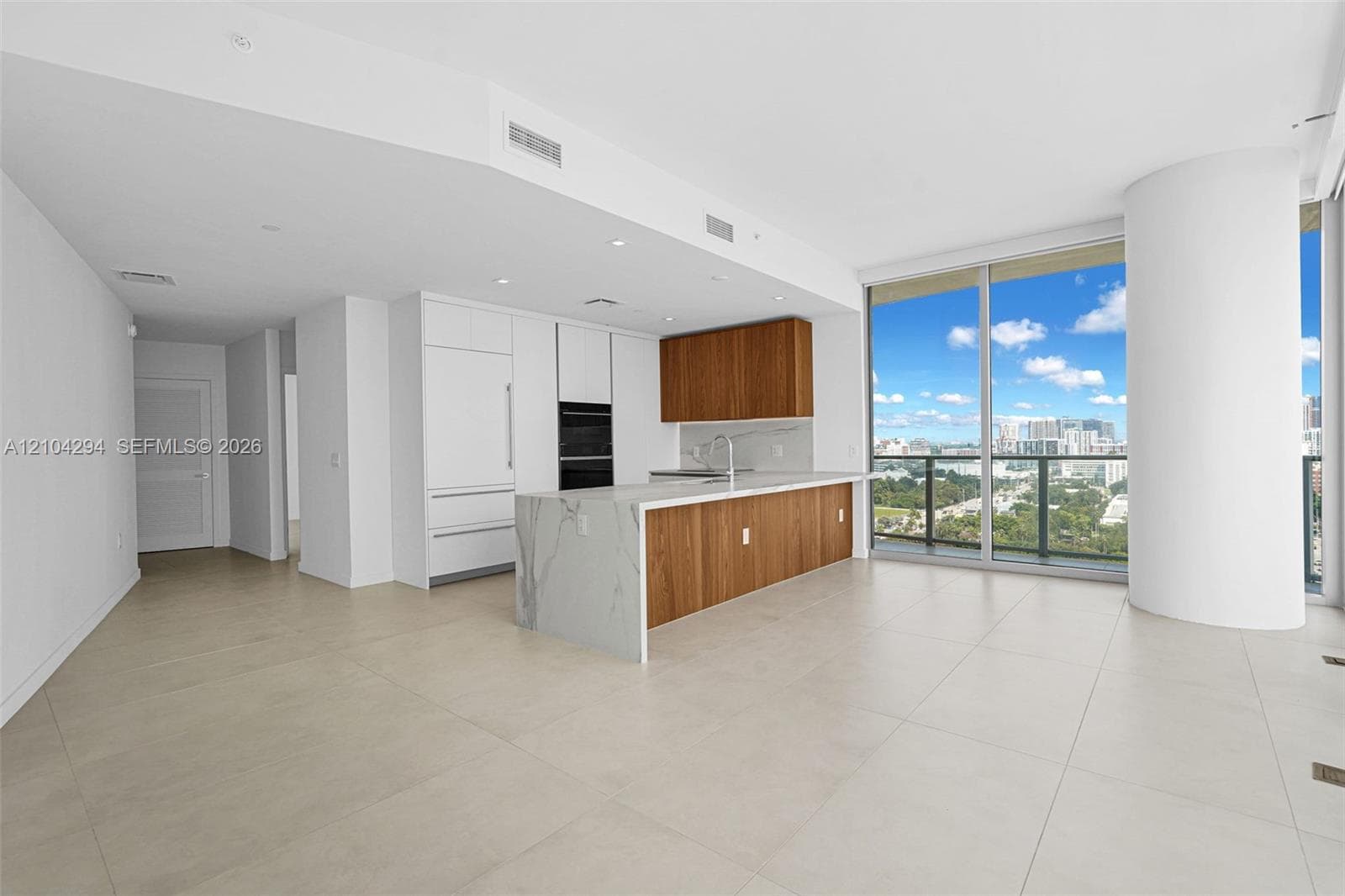 Open concept living area with light-toned tile flooring, floor-to-ceiling windows, and a modern kitchen featuring wood-finish cabinetry, integrated appliances, and a waterfall island