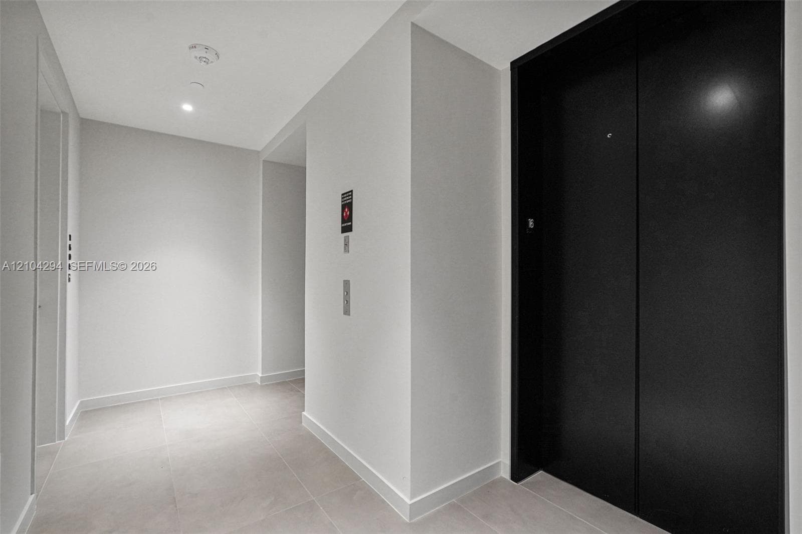 Recessed ceiling lighting illuminates the corridor, featuring a black elevator entrance, light-toned wall finishes, and square tile flooring