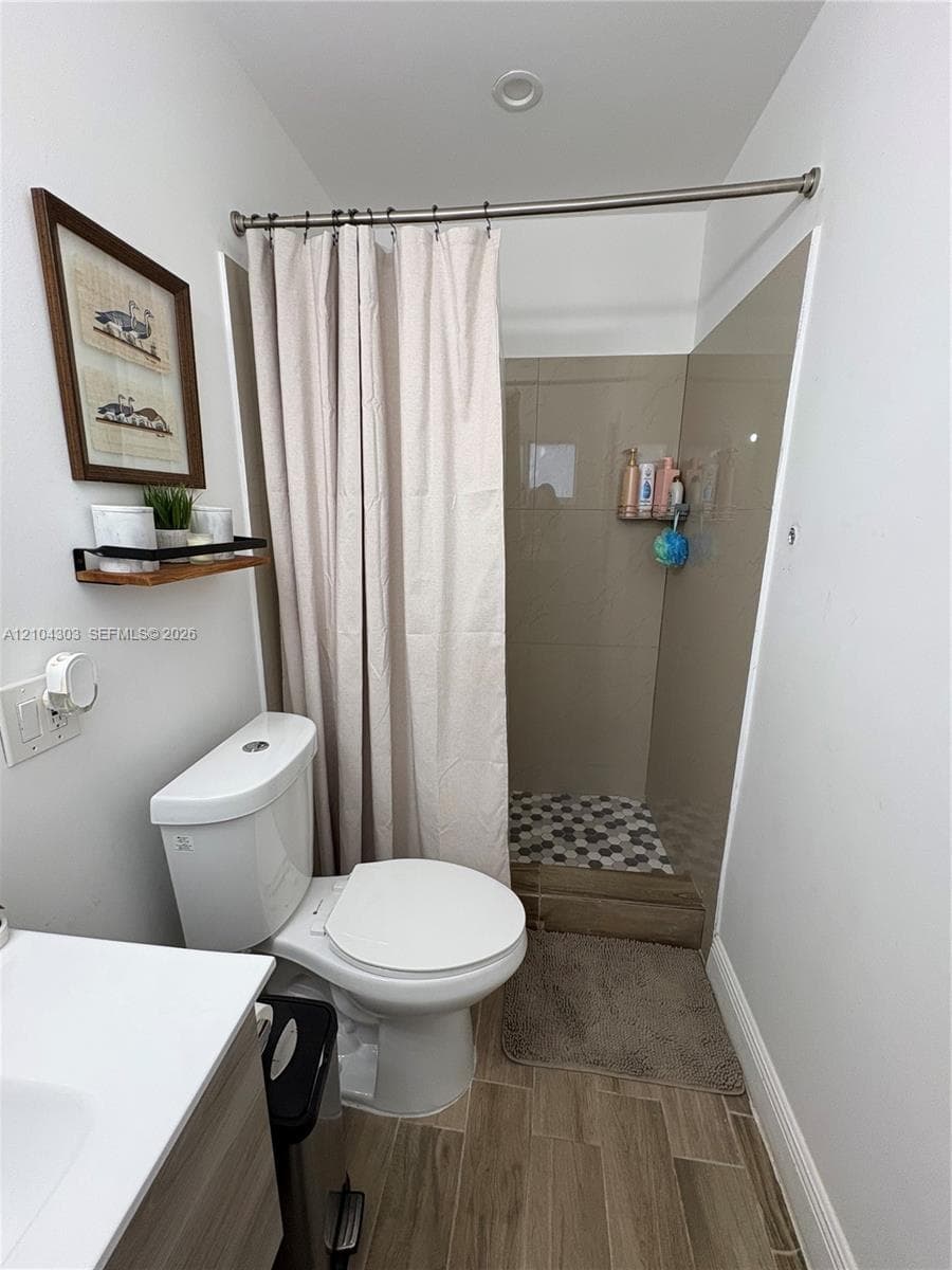 Bathroom featuring a wood-finish vanity, solid-surface countertop, walk-in shower with large-format tile surround, hexagonal tile floor inlay, and wood-finish flooring