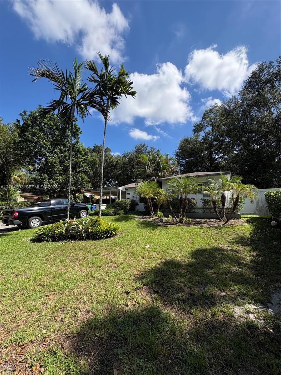 Exterior lot featuring a manicured lawn, established palm trees, low-lying shrubbery, and mature perimeter trees