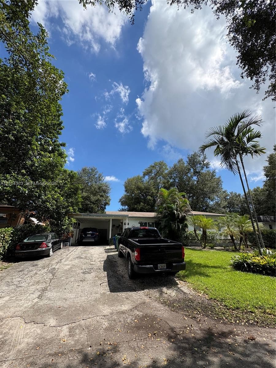 Single-story residence featuring a covered carport, expansive concrete driveway, mature landscaping, and established trees