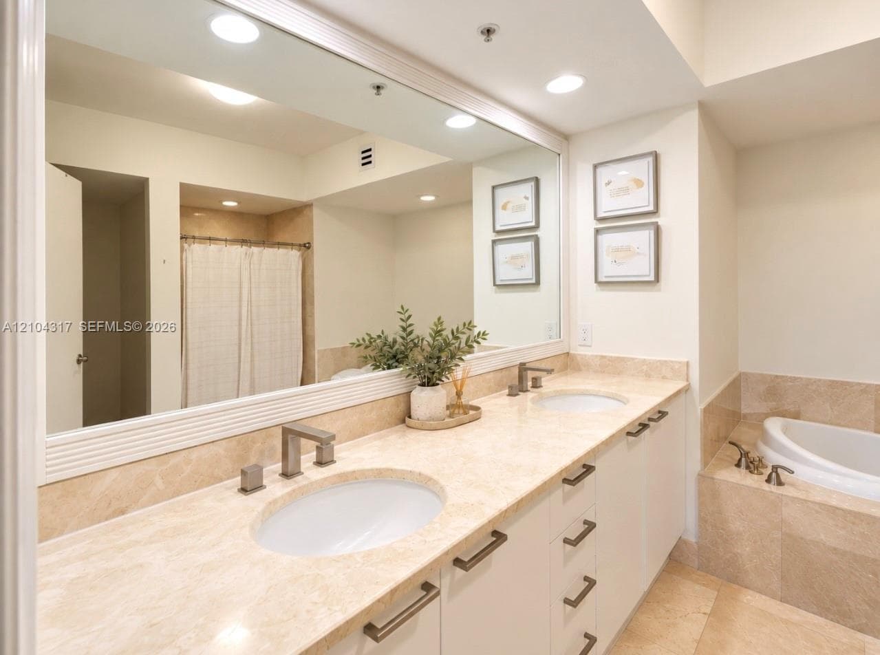 Dual-sink vanity with stone countertop, large-format mirror, recessed lighting, and built-in soaking tub with stone tile surround