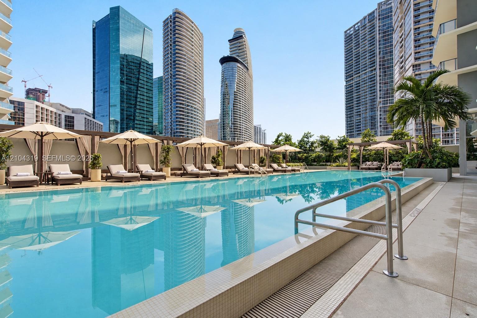 Outdoor swimming pool featuring a tiled edge, metallic handrails, and a surrounding paved deck with shaded cabanas and mature landscaping