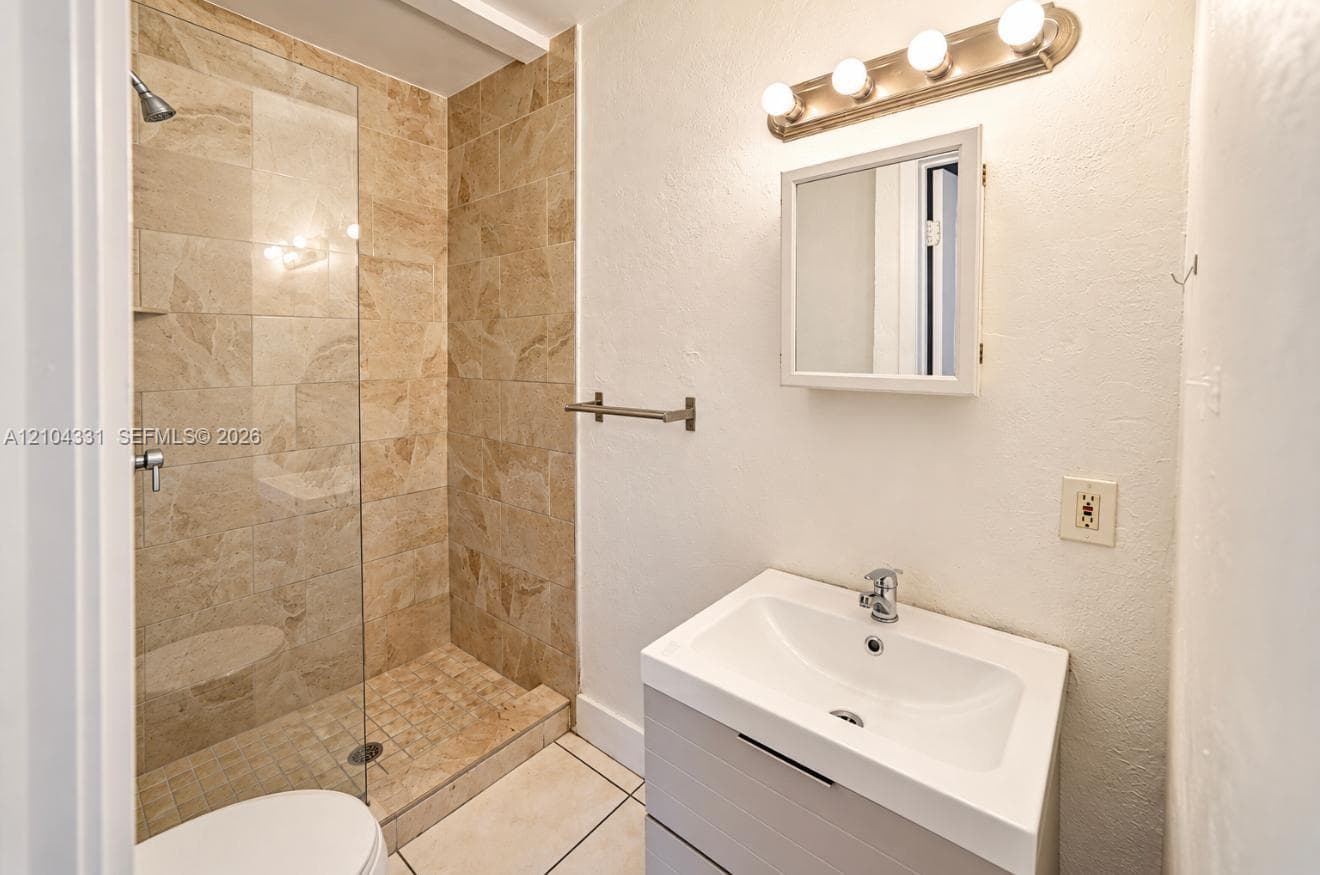 Walk-in shower featuring floor-to-ceiling stone-finish wall tiles, stone flooring, and a frameless glass door, complemented by a white vanity with an integrated sink and a four-bulb wall-mounted light fixture