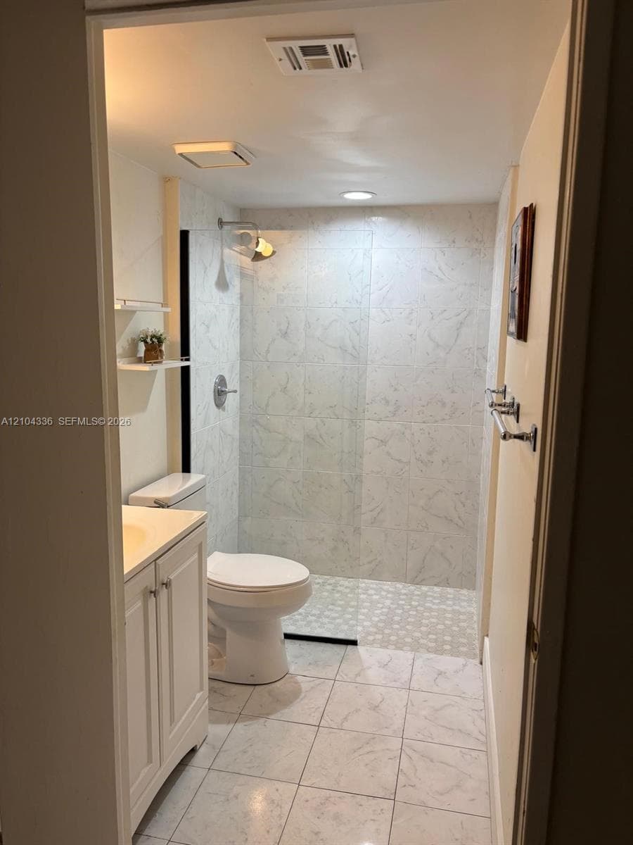 Bathroom featuring a walk-in shower with a glass panel, light-toned stone-patterned wall and floor tile, white vanity cabinet with a neutral countertop, and polished chrome hardware