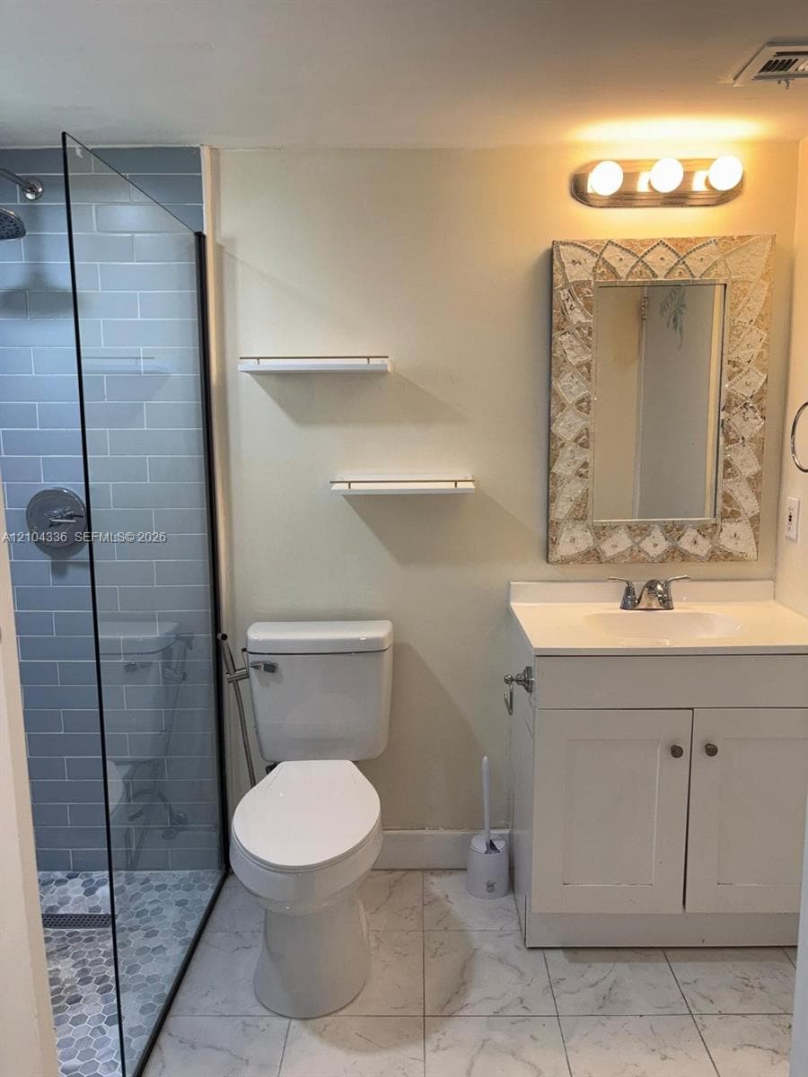 Walk-in shower with glass panel, gray subway tile wall, pebble-texture floor, white vanity with integrated basin, and two mounted floating shelves