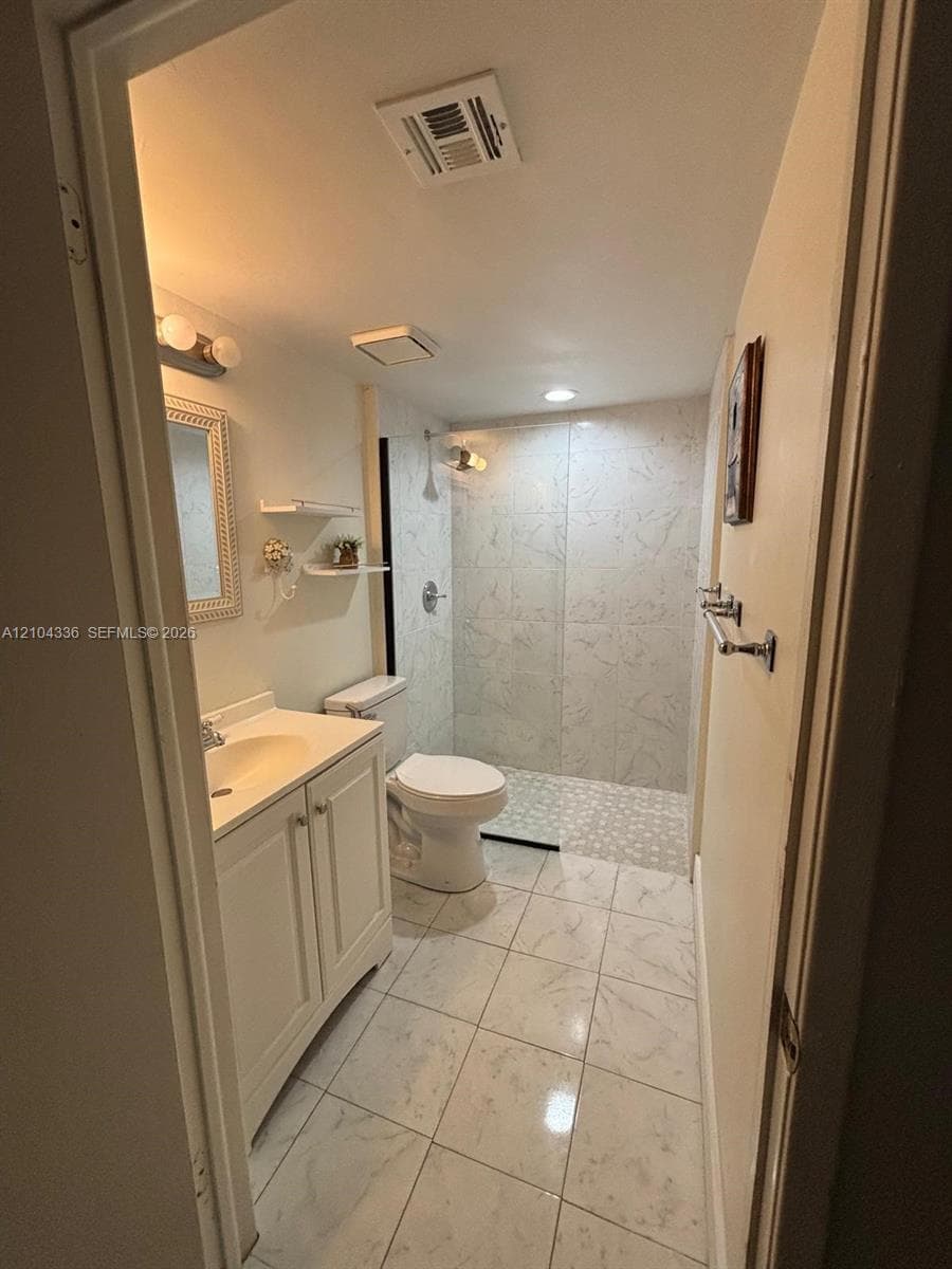 Bathroom featuring a vanity with integrated sink, walk-in shower with tiled surround, light-toned stone-finish floor tile, and wall-mounted towel bar