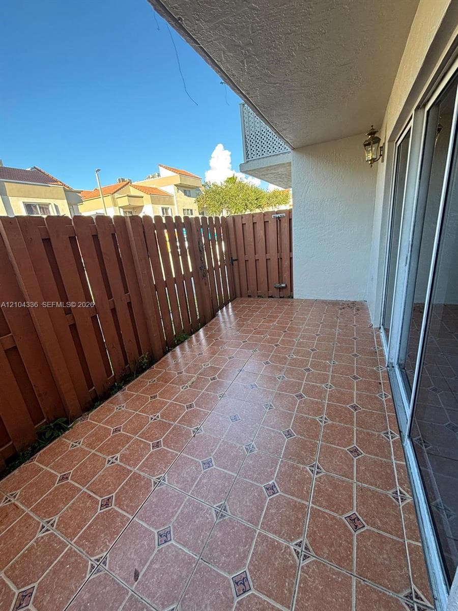 Covered outdoor patio featuring patterned tile flooring, wood-style privacy fencing, and exterior wall-mounted lighting