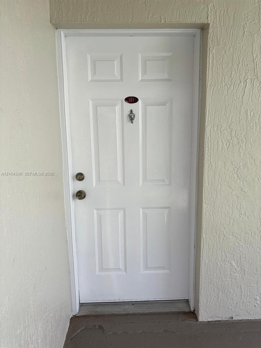 White six-panel entry door featuring a polished metal door knocker, dual-locking hardware, and a recessed number plate