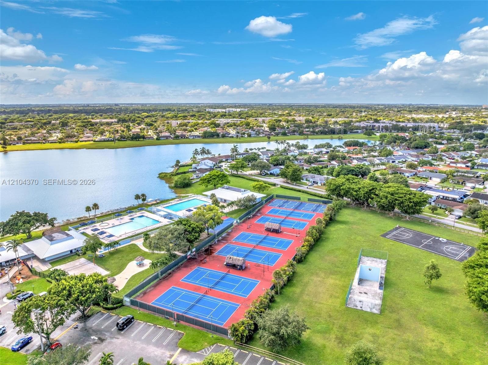 Aerial perspective of a residential lot featuring a multi-gabled shingle roof, paved driveway, expansive lawn, mature perimeter trees, and integrated landscaping