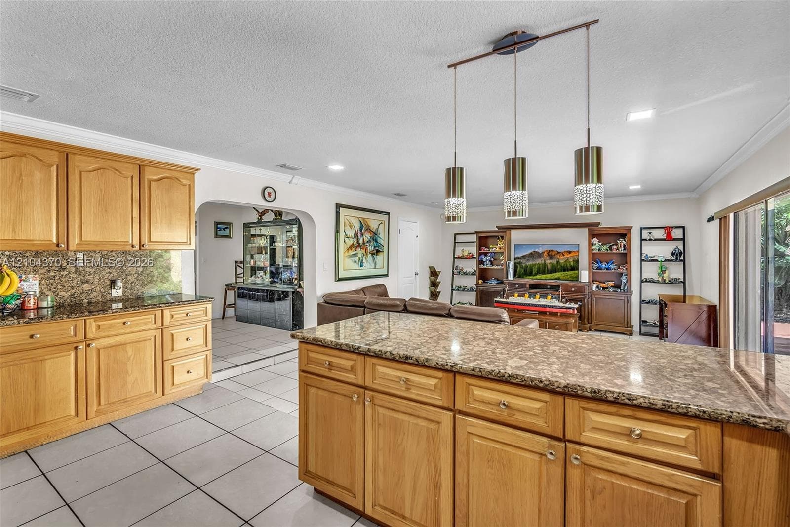 Kitchen featuring wood cabinetry, stone countertops, a tile backsplash, and a double-basin sink with a matte black faucet, illuminated by recessed lighting and a three-light pendant fixture over the peninsula