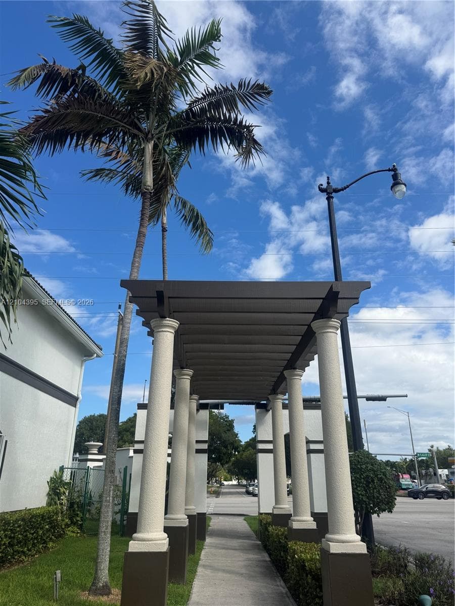 Covered walkway featuring a pergola structure supported by concrete columns, accented by mature palm trees and manicured shrubbery