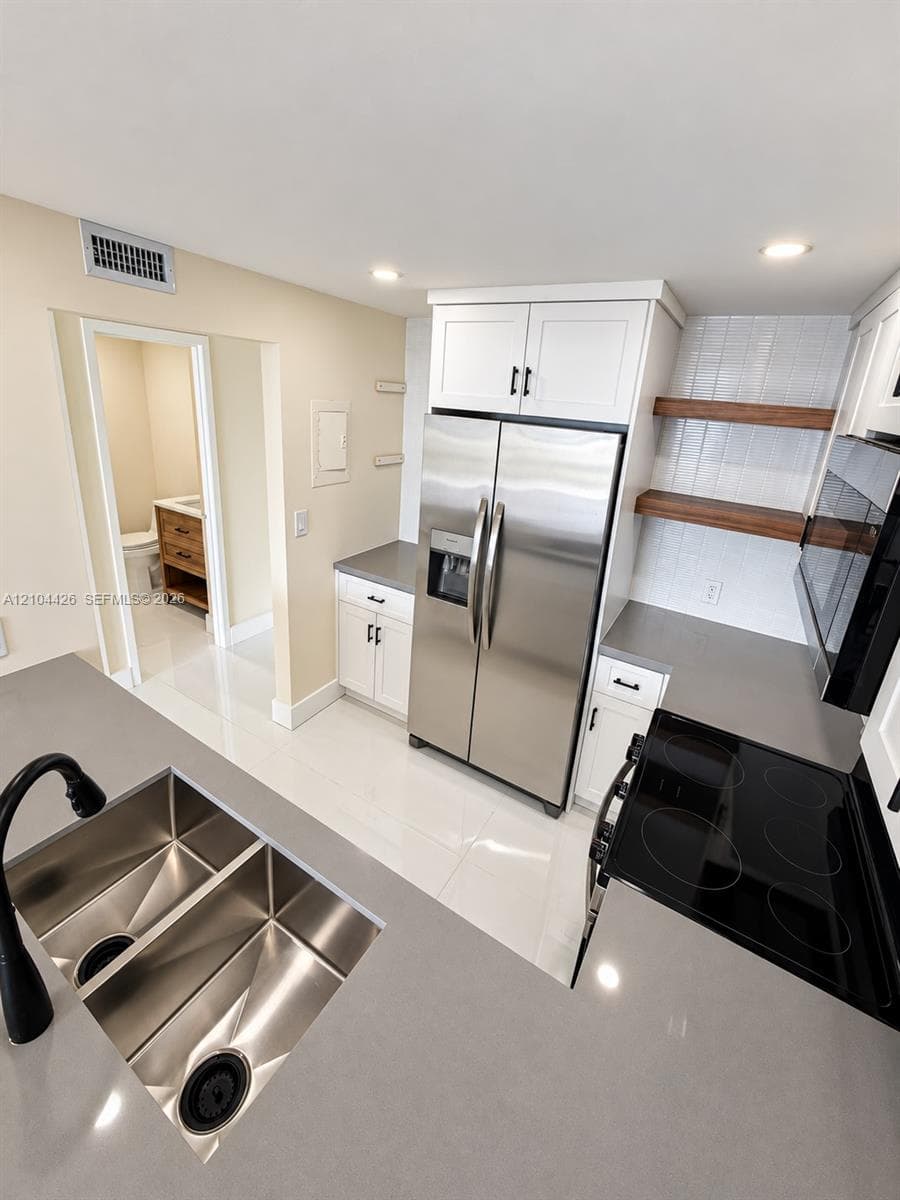 Kitchen featuring white shaker-style cabinetry, solid-surface countertops, stainless steel appliances, open wood shelving, and a double-basin undermount sink with a matte black faucet