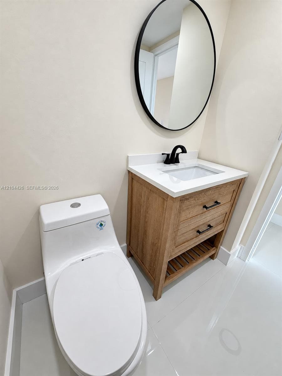 Wood-finish vanity with two drawers, open lower shelving, and a rectangular integrated sink with a matte black faucet, topped by a circular mirror with a matching black frame and a white, dual-flush, one-piece toilet