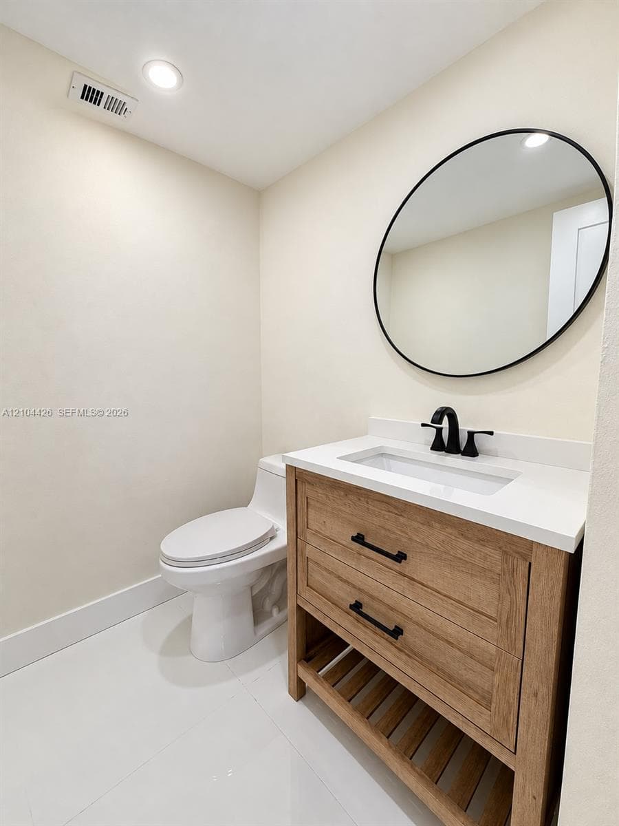 Wood-finish vanity with a rectangular sink, white countertop, matte black hardware, and an integrated slatted lower shelf, paired with a round mirror featuring a matte black frame