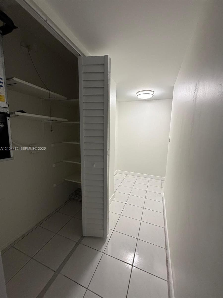 Storage space featuring white tile flooring, ceiling-mounted flush lighting, and built-in shelving enclosed by bi-fold louvered doors