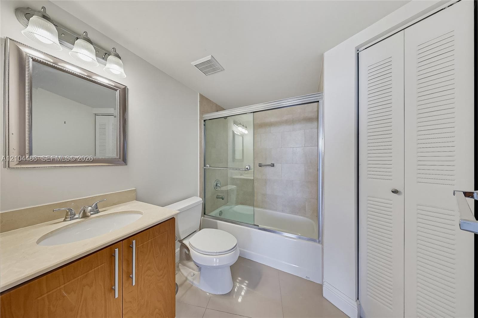 Bathroom featuring a vanity with a light-toned countertop, undermount sink, and brushed metal hardware, complemented by a tri-light fixture, framed wall mirror, glass-enclosed shower and tub combination, and bi-fold louvered closet doors