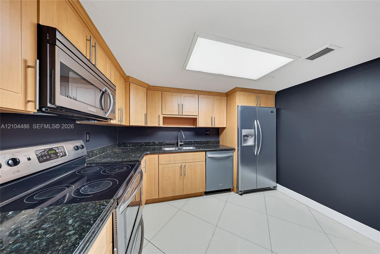 Kitchen featuring light wood cabinetry, dark stone countertops, stainless steel appliances, and large-format tile flooring