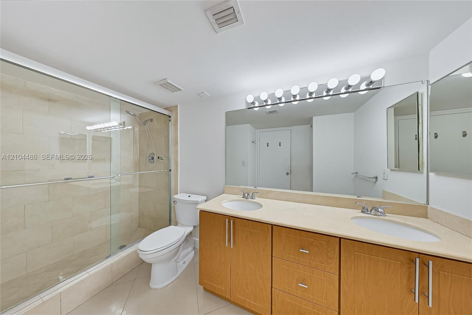 Bathroom featuring a double-sink vanity with light-tone cabinetry, solid-surface countertop, full-width vanity mirror, and walk-in shower with tile surround and glass enclosure