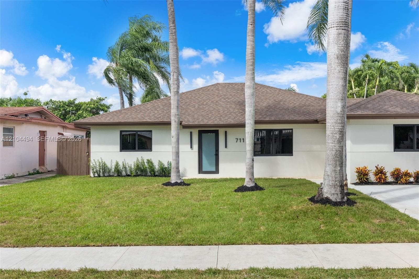 Exterior featuring a light-colored stucco facade, a hipped roof with asphalt shingles, framed windows with dark trim, a glass-panel entry door, and manicured landscaping with mature palm trees