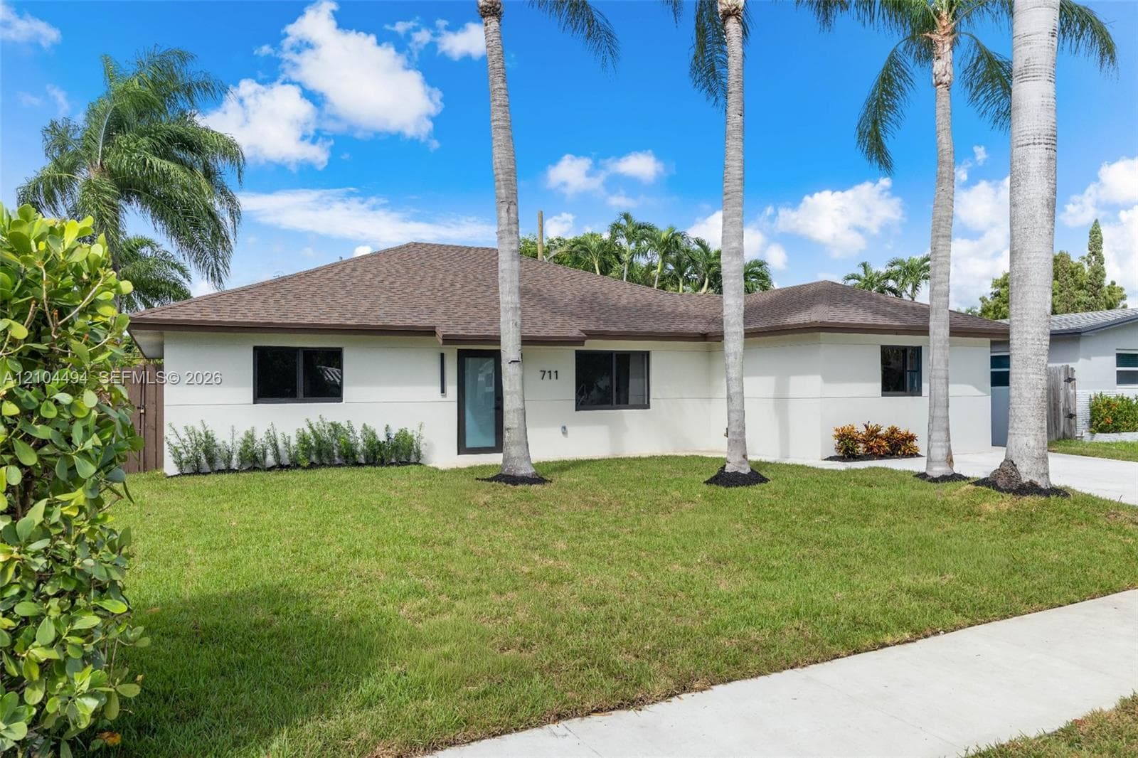 Single-story exterior featuring a light-toned stucco façade, a hip roof, dark-framed windows, and mature palm trees