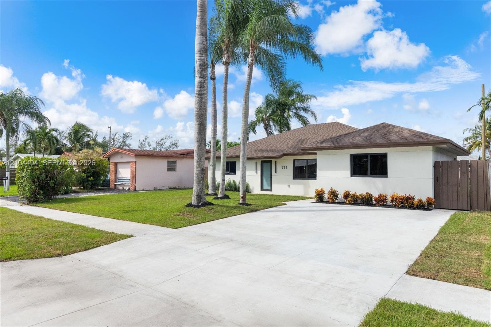 Single-story residence featuring a neutral stucco exterior, a shingled hip roof, a concrete driveway, mature palm trees, and a wooden privacy gate