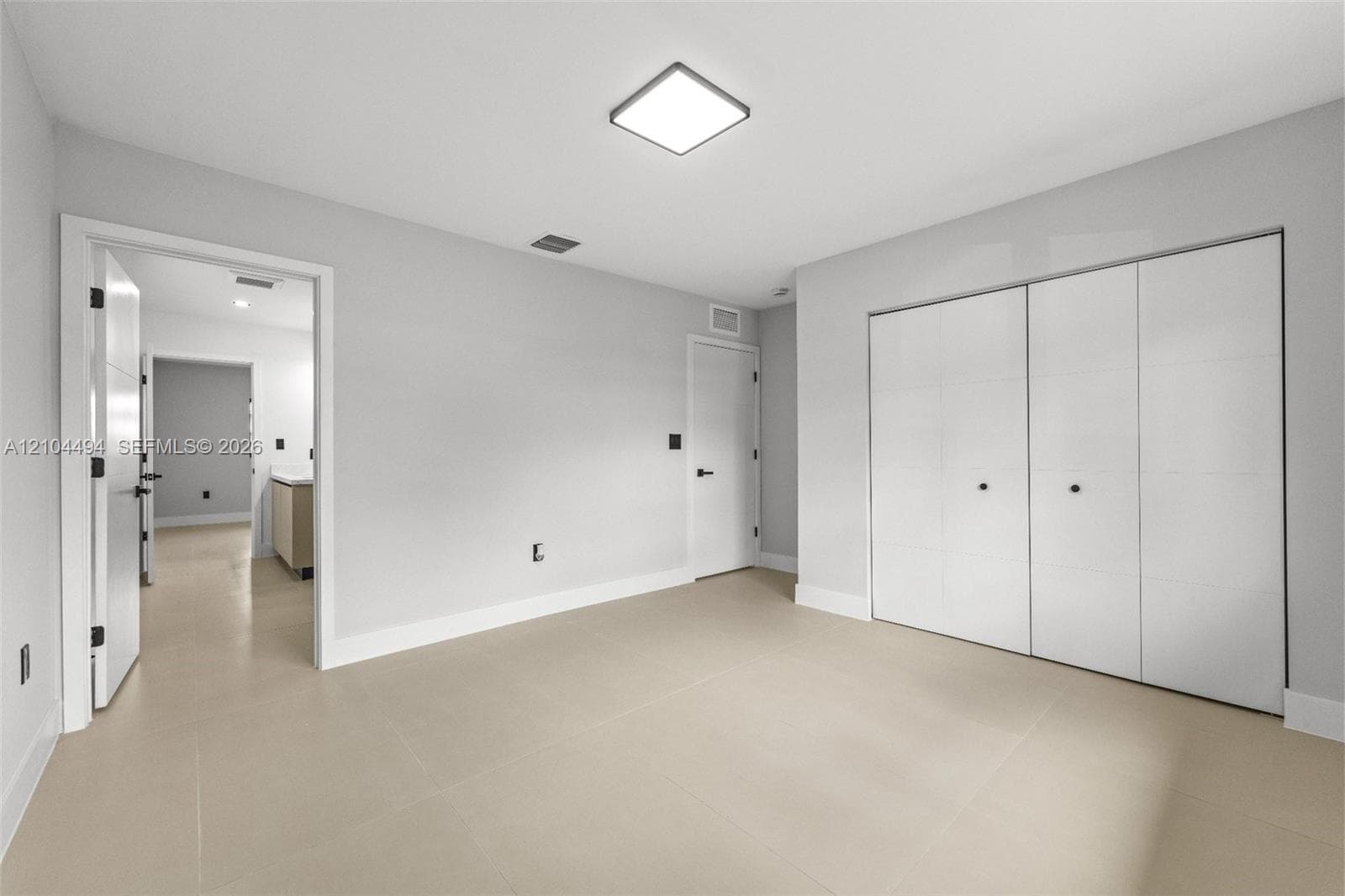 Neutral-toned interior space featuring light-colored tile flooring, flush-mount square ceiling lighting, and a multi-panel sliding closet system with matte black hardware
