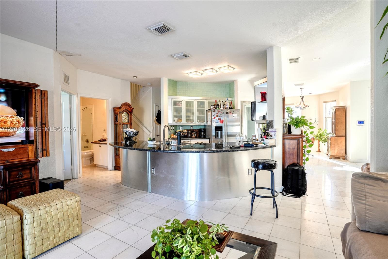 Curved kitchen island featuring a dark stone countertop and stainless steel facade, light-toned tile flooring, recessed ceiling lighting, and white cabinetry with glass-front uppers