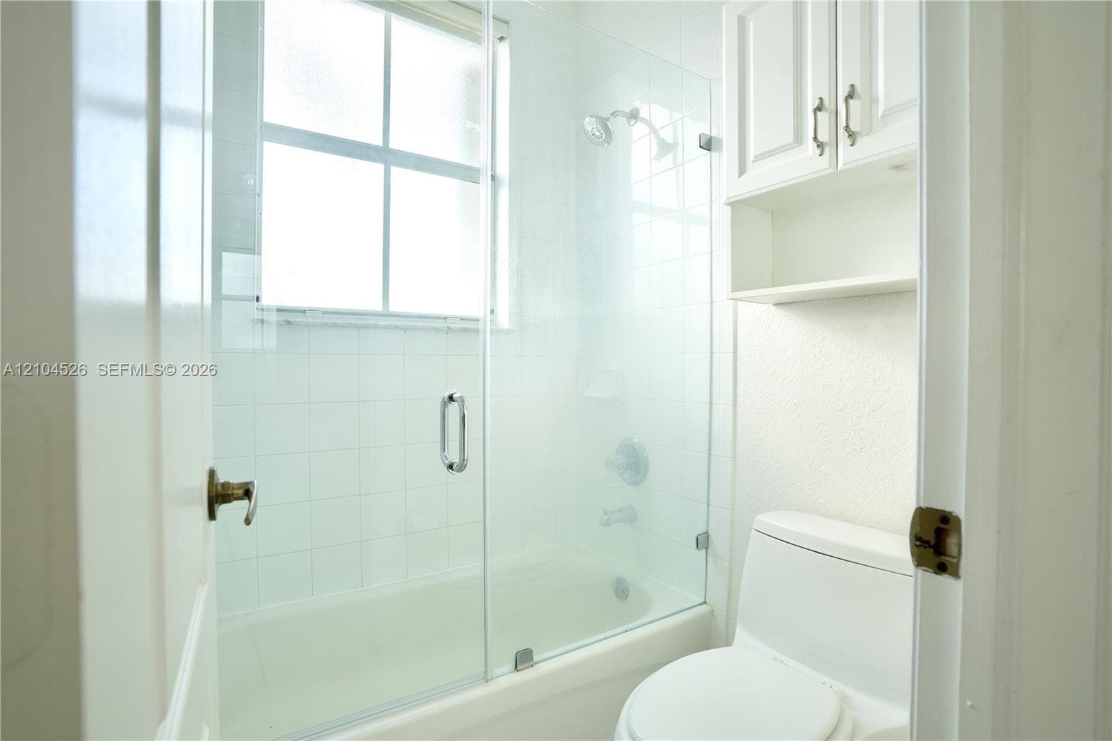 Bathroom featuring a tub-shower combination with a glass door enclosure, white tile surround, integrated window, and built-in upper cabinetry