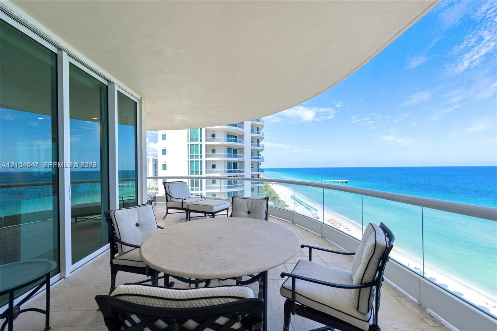 Curved covered balcony with tile flooring and a frameless glass railing, featuring floor-to-ceiling glass doors and coastal scenery