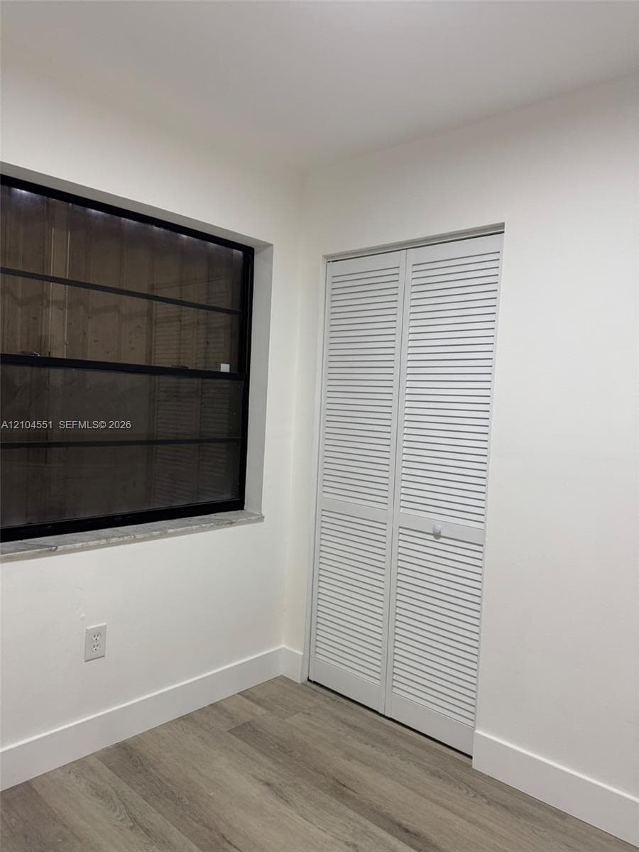 Room featuring wood-finish flooring, white louvered closet doors, and a window with a black frame and stone sill