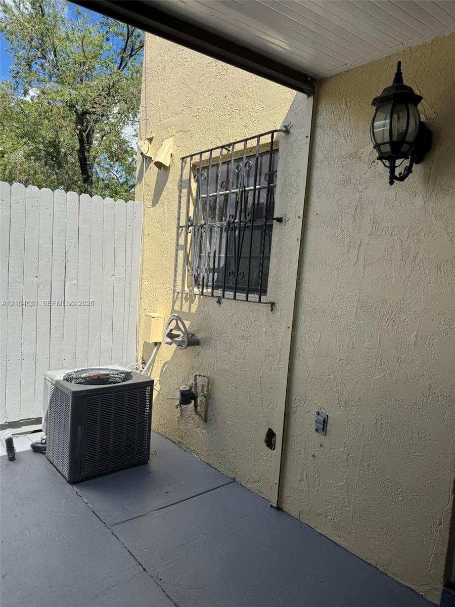 Covered patio featuring a textured stucco exterior, metal window security bars, a wall-mounted lantern-style exterior light fixture, and a vertical wood privacy fence