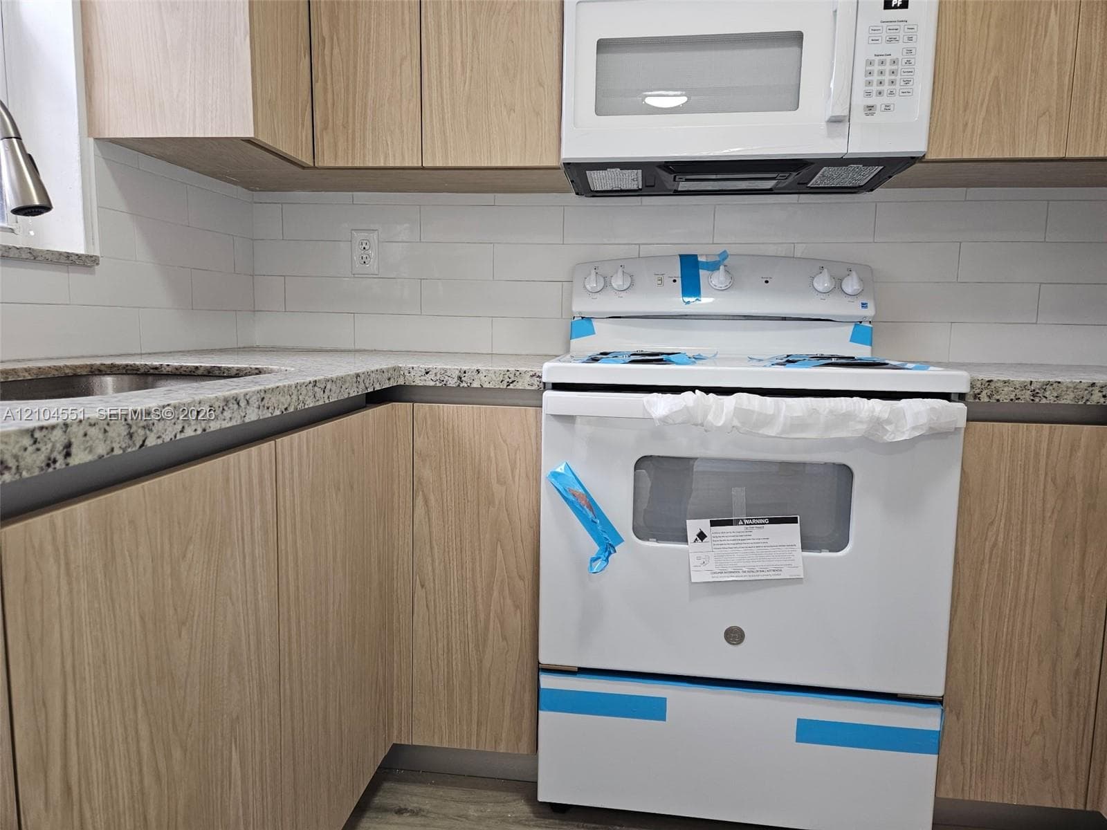 Kitchen featuring flat-panel wood-finish cabinetry, speckled stone-finish countertops, white subway tile backsplash, and white appliances