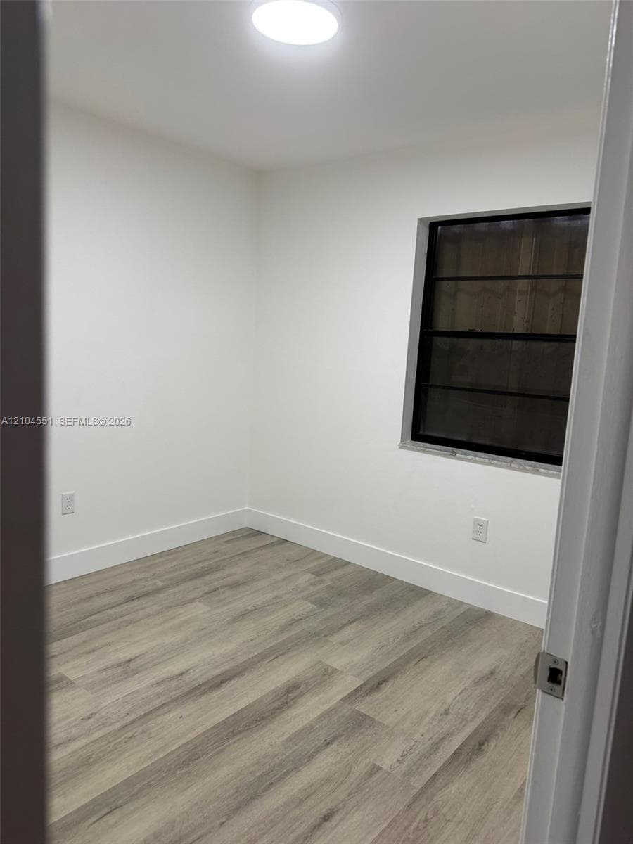 Neutral-toned interior space featuring wood-finish flooring, white walls, a flush-mount ceiling light fixture, and a single window with a dark frame