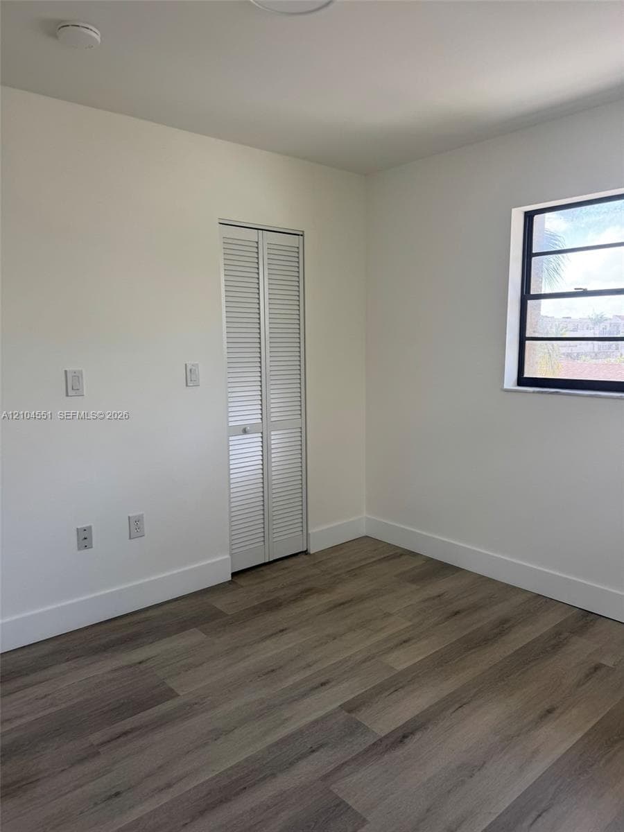 Room featuring wood-finish flooring, white walls, louvered closet doors, and a window with a black frame
