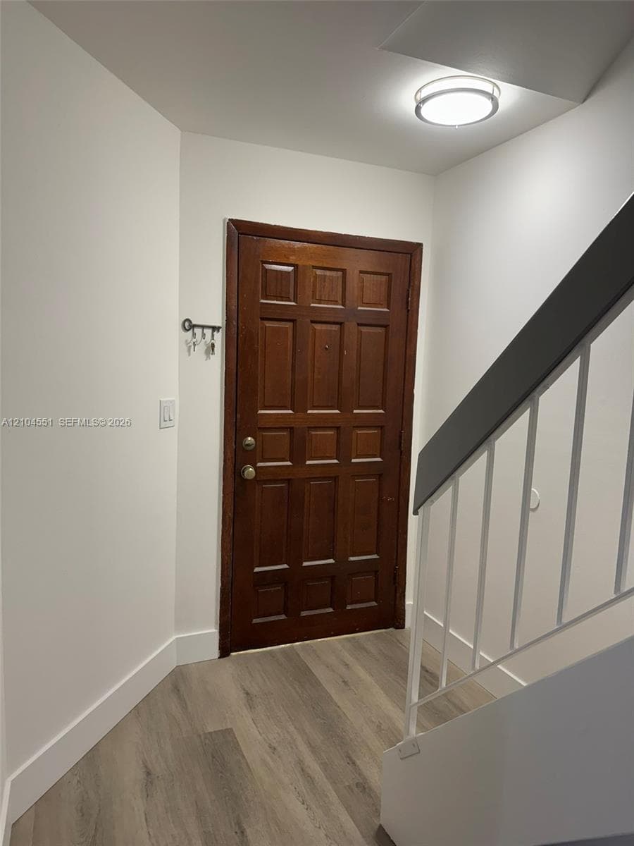 Entryway featuring a solid wood-finish door with multi-panel detailing, neutral wood-finish flooring, and a staircase with a white railing and dark-toned handrail, illuminated by a flush-mount circular ceiling fixture