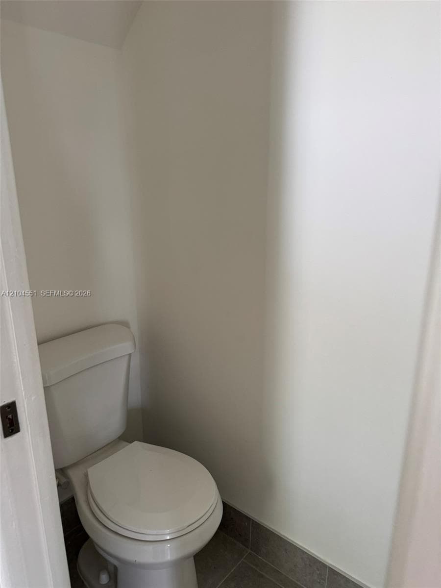 Enclosed space featuring a white porcelain toilet and neutral-toned tile flooring