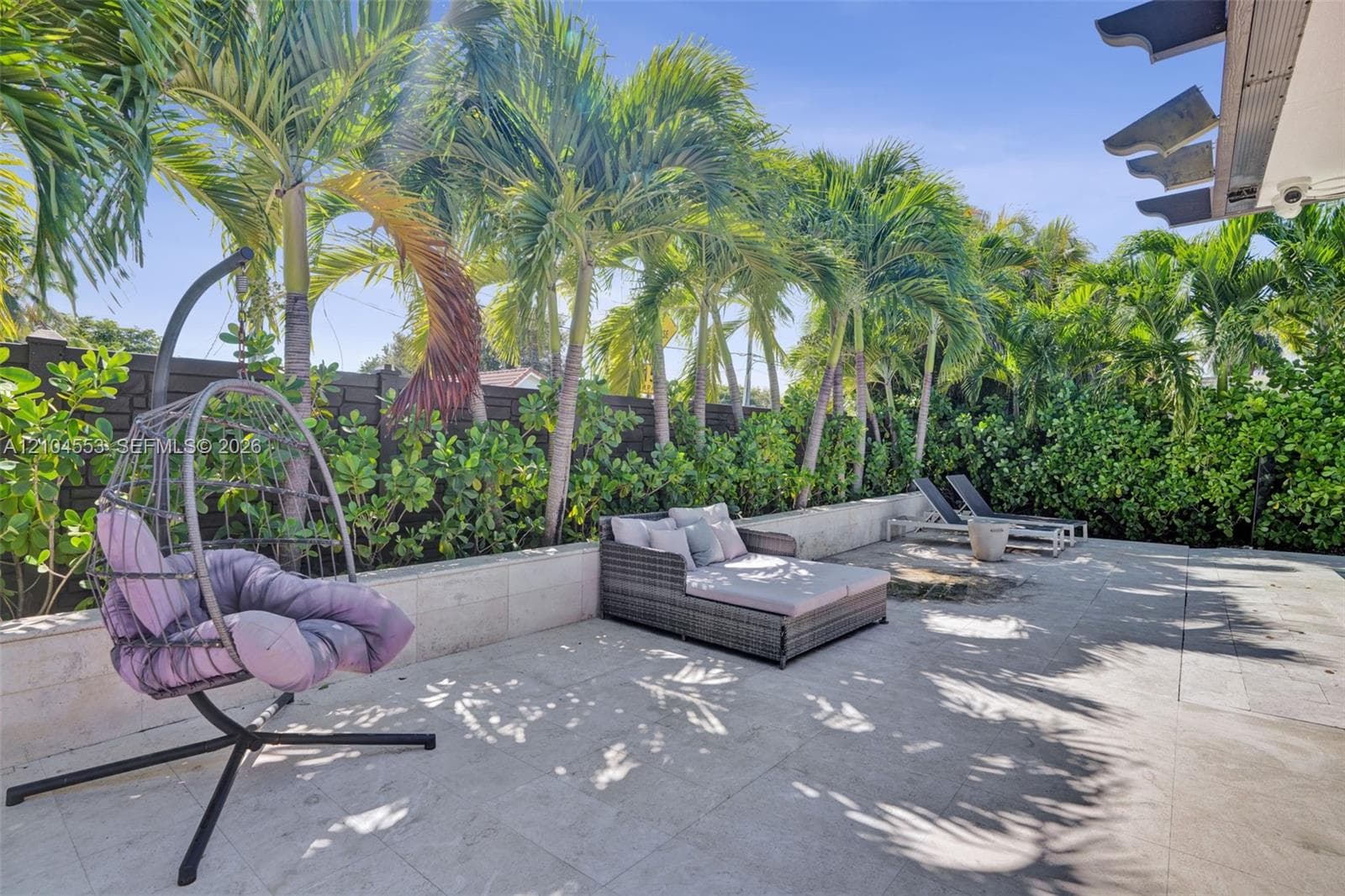 Rear patio featuring stone-tile flooring, a built-in stone retaining wall, mature palm trees, and perimeter privacy fencing