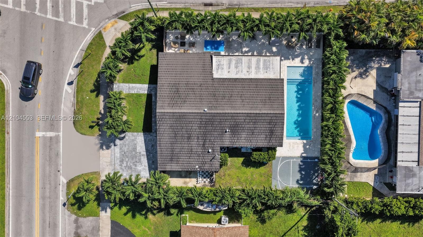 Aerial perspective of a fenced property featuring a rectangular swimming pool, concrete patio, basketball court, and perimeter landscaping