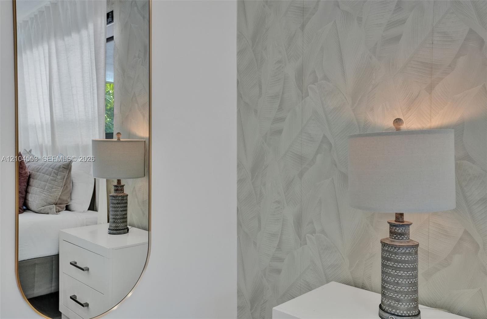Full-length arched wall mirror with a slender gold-tone frame, white two-drawer built-in cabinetry, and textured botanical-patterned wall covering