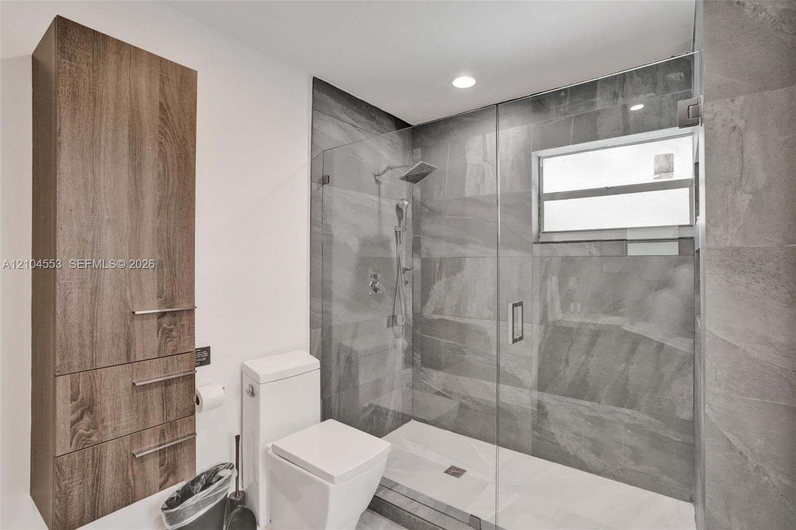 Walk-in shower featuring glass enclosure, large-format grey tile surround, and rainfall showerhead, accompanied by tall wood-finish cabinetry and a white high-profile toilet