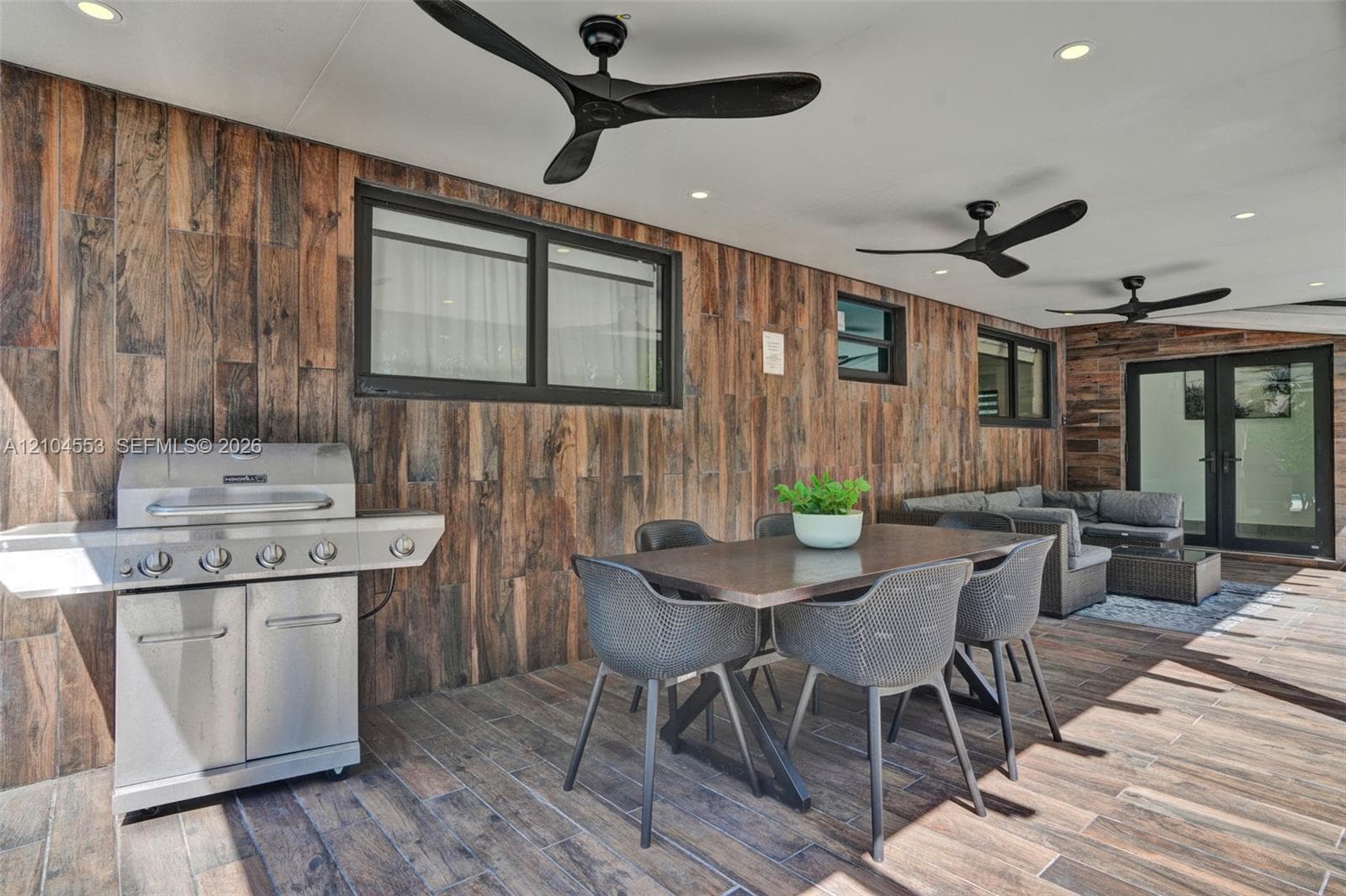 Covered patio featuring wood-finish tile wall cladding, wood-finish floor tiling, a built-in stainless steel grill, recessed lighting, and three matte black ceiling fans