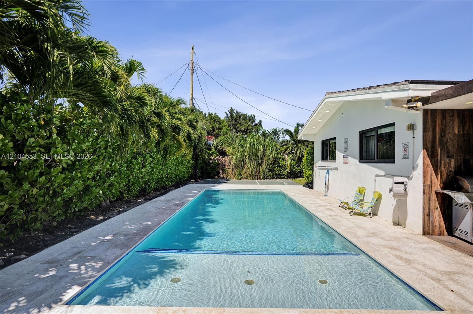 Rectangular in-ground pool with tile trim, expansive light-toned stone patio, exterior wall with wood-finish paneling, and established perimeter hedging