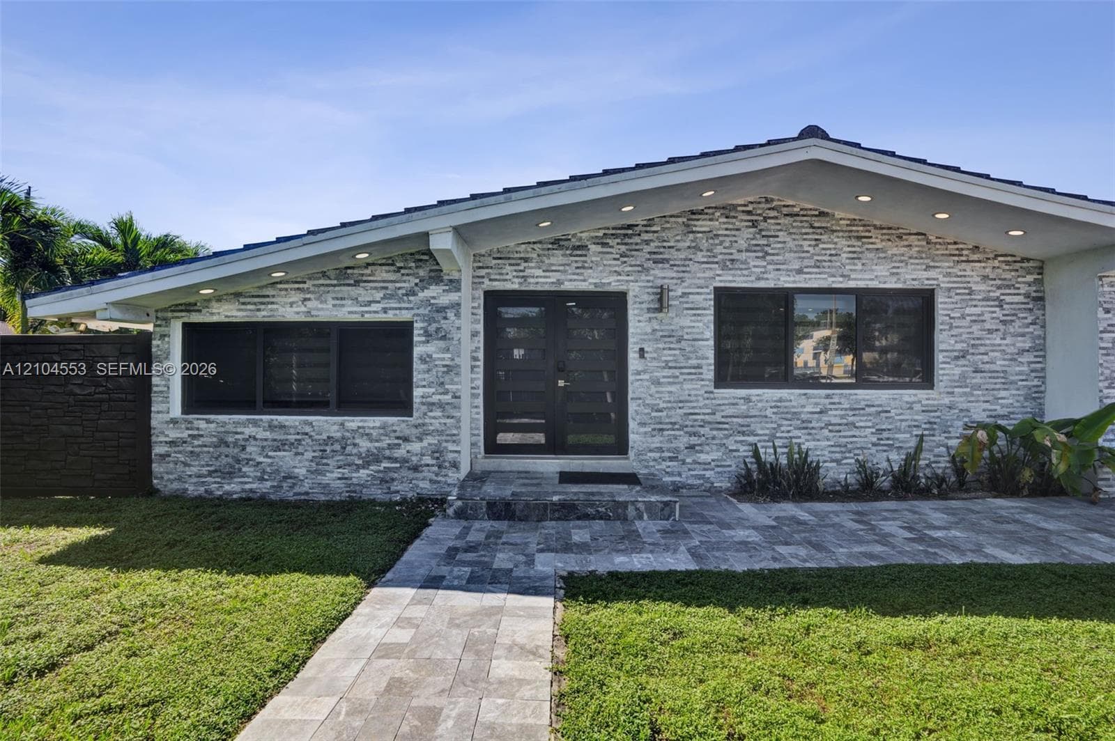 Exterior facade featuring a stacked stone veneer, double glass-panel entry doors, a low-pitch roofline with recessed soffit lighting, and a paved stone walkway